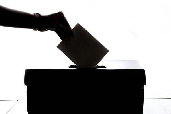 Fotografie einer Hand die einen Umschlag in eine Wahlurne wirft.